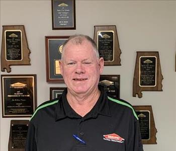 Photo of male employee smiling in black collared shirt with SERVPRO logo in front of wall of awards